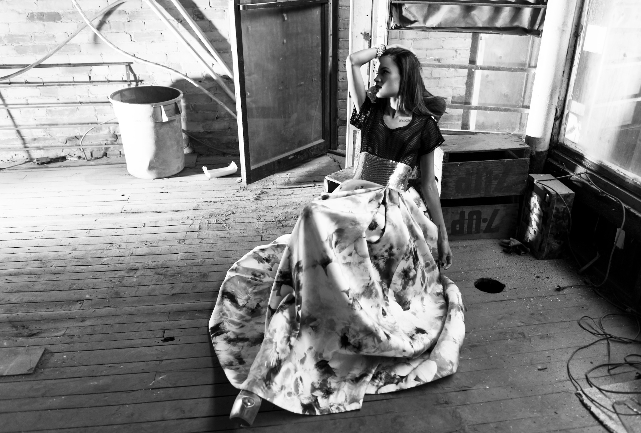 Black-and-white ALLWOOD editorial photograph of a seated model in a sweeping patterned skirt, surrounded by weathered windows and wooden floorboards