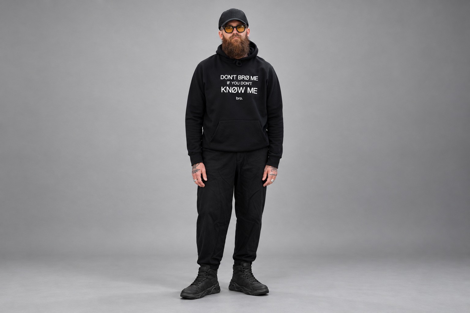 Studio styling visual of a bearded model in the black graphic hoodie, ALLWOOD amber aviators and black high-top sneakers against grey seamless paper
